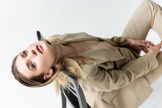 trendy young woman in trench coat and scarf sitting on stool while posing on grey (foto: Shutterstock)