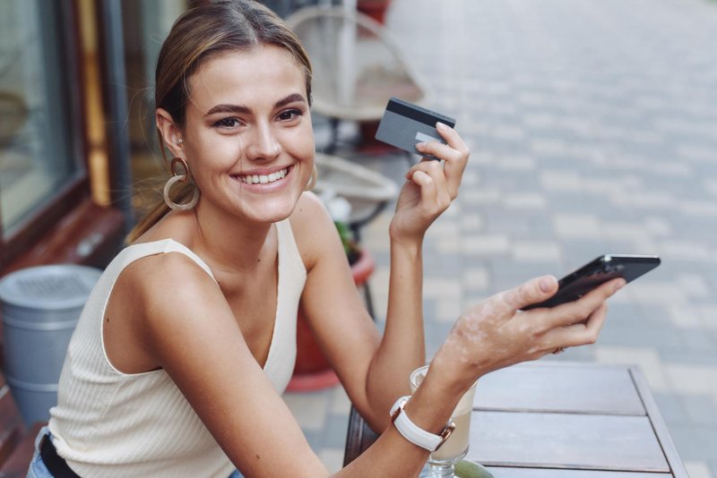 Smiling woman holding phone and credit card outdoors in cafe. A beautiful model looks at the camera and pays for purchases with a card and phone (foto: Shutterstock)